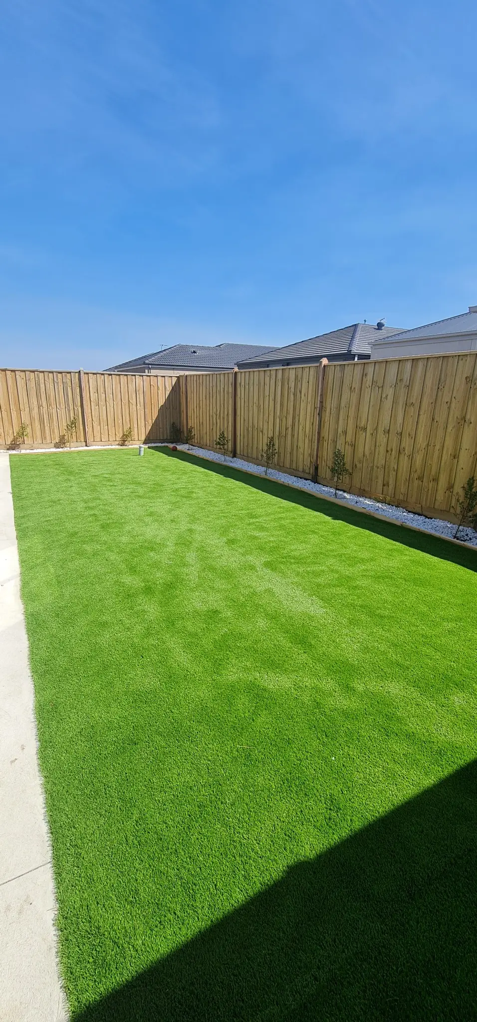 Completed artificial turf installation with timber fencing, white pebbles and young boundary planting
